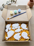 A cardboard box filled with yellow shredded paper holds six white fish-shaped items, with three small paint pots, a paintbrush, and decorative eucalyptus branches on a light wood surface, its the diy pain kit.