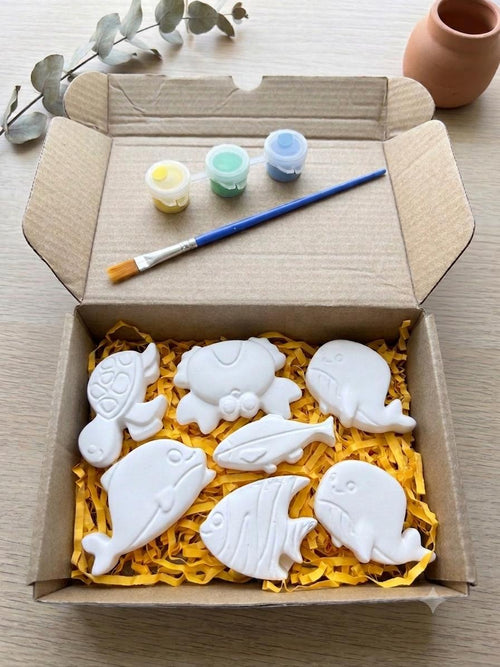 A cardboard box filled with yellow shredded paper holds six white fish-shaped items, with three small paint pots, a paintbrush, and decorative eucalyptus branches on a light wood surface, its the diy pain kit.